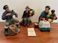 Photo of three Royal Doulton figurines on a wooden surface, showing all three characters sitting and holding balloons or a lobster, with a MaxSold identification card in front.