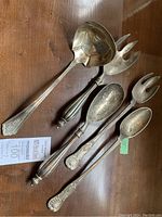 Five silver plated serving utensils on a wooden surface with a MaxSold tag. Utensils include spoons and forks with ornate handles.
