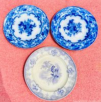 Three antique plates placed on a pink surface; two flow blue plates with blue floral patterns, and one mulberry plate with bird design.