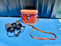 Vintage Bell and Howell binoculars placed beside brown leather carrying case with shoulder strap, showing the full set.