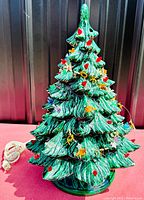 Full view of vintage ceramic Christmas tree showing green textured foliage with red bulbs and tinsel garlands, electrical cord off to the side.