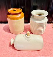 Photo showing the set of three stoneware items: a lidded pickle jar, ribbed vase, and hot water bottle with cork stopper all on a pink cloth background.