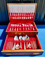 Wide view of entire wooden chest open showing two tiers and a drawer filled with silverplate flatware, arranged in rows on red velvet lining.