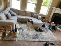 Overview of a large beige sectional sofa with the 8x10 foot area rug taped underneath, showing some clutter around on the rug and carpet floor.