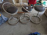 Three metal swivel bar stools with black seats and circular metal bases on concrete floor, showing overall condition including rust and scratches.
