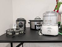 Lot items displayed on black table: Sunbeam Crock-Pot, Aroma rice cooker, Cuisinart frozen yogurt-ice cream maker, and circular tea light holder