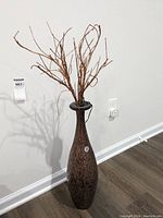 Full view of tall brown Dale Tiffany vase on floor against wall, holding multiple twigs and branches with string lights.