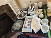 Photo of assortment of white ceramic appetizer spoons, various white ceramic serving plates with embossed design, clear etched glass plate, large clear glass mixing bowl, black and white patterned small rectangular dish and napkins on black table.