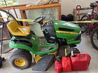 Front-left perspective of green and yellow John Deere LA115 riding mower parked indoors with three red gas cans in front.