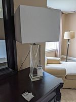 Full view of the glass based table lamp with white rectangular shade on black table near a mirror and beige furniture