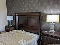 View of one table lamp on wooden nightstand next to bed and wallpapered wall, showing lamp in context and full height including shade.