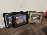 Three prints shown with two New York City themed prints framed in black and one floral print framed in gold ornate frame with green mat, one NYC print unframed and leaning behind others.