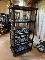 Front and side view of the five level black plastic shelving unit showing grid shelves and cylindrical legs in basement setting.