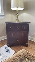 Front view of dark wood side table chest of drawers with 3 drawers, metal ring pulls, and metal corner guards. Lamp on top.