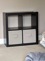 Front view of black wood grain cubby shelf with two beige fabric storage bins in lower compartments, placed against a beige wall on carpeted floor.