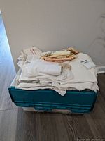 Pile of folded linen and embroidered table linens on top of blue plastic storage crate, including folded tablecloths and napkins.
