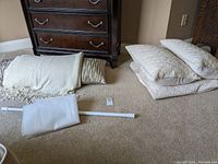 Quilted bedding items including pillows, folded blanket or duvet, and an extension rod with shower curtain lying on carpet near wooden furniture.