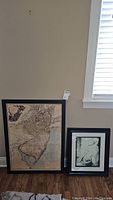Both framed prints leaning against wall: large 1778 New Jersey map print in dark frame and smaller grayscale woman's back print in wooden frame.