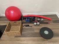 Wide view of all exercise equipment including yoga ball, balance disk, dumbbells, body bar, pool noodle, and basket on wooden floor.
