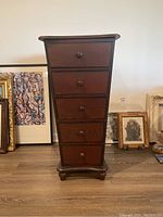 Front view of wooden lingerie chest showing five drawers and wooden knobs, with artworks leaning against the wall behind it.