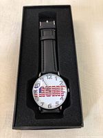 Black wristwatch with leather straps in a black box. White dial with 'TRUMP' written in American flag pattern, pin pulled.