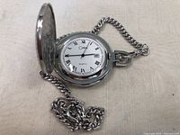 Open pocket watch showing white dial with Roman numerals and black hands attached to silver chain.