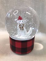 Front view of the snow globe showing the white Golden Retriever figurine inside a glass dome and the red and black plaid base.