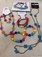 Top-down photo of assorted colorful kids jewelry on a beige surface including bracelets, necklace with butterfly pendants, and a decorative hair accessory.