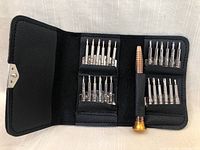Open black textured synthetic leather case displaying 24 metal precision screwdriver bits neatly organized in elastic slots, and a copper-colored mini screwdriver handle with a gold base in the middle.