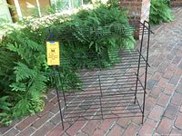 Front view of black wire metal shelving unit showing 4 wire tiers with rust and some wear, placed on brick flooring with plants in background.