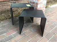 Pair of black metal nesting tables displayed outdoors on brick ground, showing overall appearance, size, and shape.