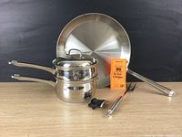 Full set of a 13-inch All Clad frying pan, Belgique 2 qt double boiler with lid, meat fork, and small peeler on a wooden surface.