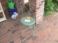 Full view of the antique soda fountain chair showing round seat, metal legs, and green chippy paint finish placed outdoors against brick wall.