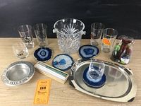 Full lot with crystal ice bucket at center, four blue geode coasters in front, clear glass tumblers and glasses placed around, red wine bottle coaster, wine pump on right, stainless steel serving tray beneath some items, and set of glass straws with cleaning brush in front left.