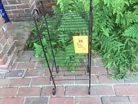 Black metal wire stand placed outdoors on brick patio beside green ferns, showing front and top view of two shelves.