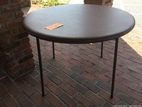 Full view of round folding table with brown padded top and metal legs on a brick floor.