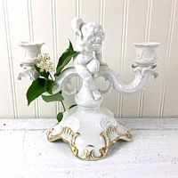 Front view of white ceramic double candleholder featuring a cherub figure with gold decoration on the base, sitting on a white surface against a white paneled backdrop, with a sprig of leaves and white flowers nearby.