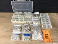 Overview photo of all plastic boxes and labeled plastic bags containing various jewelry findings such as earwires, clasps, and jump rings arranged on a table surface.