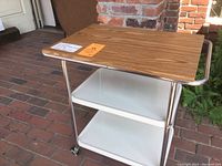 Front-angle view of vintage kitchen utility cart showing wood laminate top, two white enamel metal shelves, chrome tubular frame, and locking wheels.