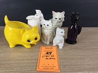 Six cat-themed items including three white cat figurines, purple art glass cat, yellow cat planter, and white cat cream pitcher with kitty handle on black background.