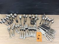 Wide view showing assortment of forks, knives, teaspoons, serving spoons and two serving utensils arranged on table with auction tag