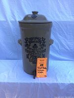 Front view of the beige salt glaze ceramic water filter with black text, molded handles, lid, and spigot, photo taken to show the entire functional and design features, as well as the size tag.