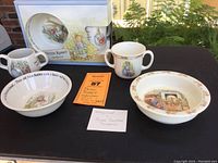 Overview photo showing the full set of Peter Rabbit Bunnykins dishes including three bowls, two two-handled cups, and one single handled cup with the original boxed set visible.