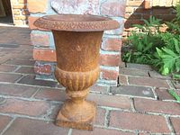 Rusty iron urn plant holder standing outdoors on brick patio near a brick wall and plants, showing overall shape, rusted surface, decorative rim, and pedestal base.