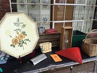 Photo shows a collection of folk art themed items including the wooden floral tray, metal grater, red funnel, basket and other decor items on a black table outdoors.