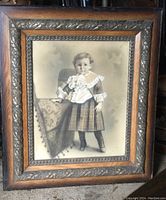 Full view of the large framed portrait showing the young boy standing beside a patterned draped table, with visible wood frame and metal decorative details.