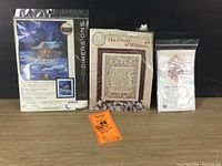 Three needle art kits displayed on a table: unopened Winter Sky Cabin needlepoint kit by Dimensions, open but complete The Prayer of St. Francis cross-stitch kit, and a sealed Alphabet Sampler kit.