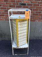 Full view of the vintage revolving metal key rack showing the rectangular white metal frame, two shelves, and the revolving central yellow and white panel with hooks.