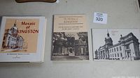 Three Kingston-related books arranged side by side: 'Mosaic of Kingston', 'The Old Stones of Kingston' and 'Kingston City Hall' booklet.