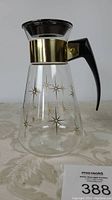 Front view of clear glass carafe with gold starburst pattern, black plastic lid and handle, gold metal band around neck.